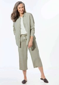 Helles grünes Leinenhemd und cropped Hose mit Kordelzugbund, kombiniert mit dunklen gewebten Slippern. Hintergrund ist schlicht weiß.