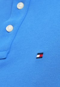 Μπλε βαμβακερό πόλο πουκάμισο με τρία λευκά κουμπιά και ένα μικρό κεντημένο λογότυπο σε κόκκινο, λευκό και μαύρο στη αριστερή πλευρά.