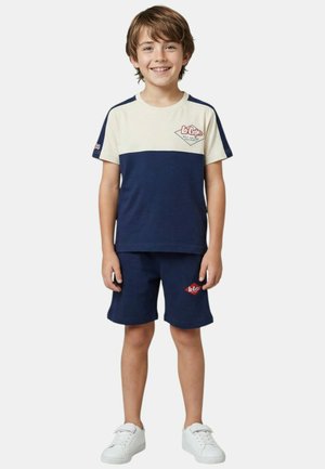 Jeune garçon souriant, portant une chemise à manches courtes bleu marine et blanche avec un short bleu marine assorti et des baskets blanches, debout devant un fond uni.