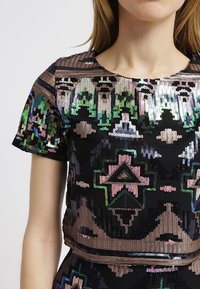 Černý krátký top s flitry s živými geometrickými vzory v zelené, růžové a modré barvě. Krátké rukávy a kulatý výstřih viditelné.