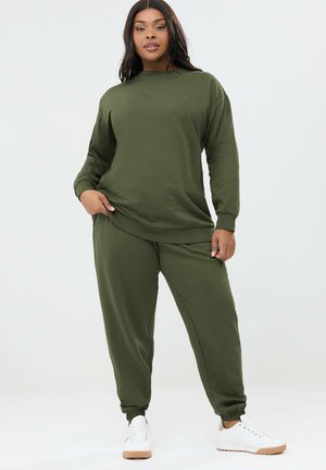 Frau trägt olivgrünes Langarm-Sweatshirt und passende Jogginghose mit weißen Turnschuhen, steht vor einem schlichten hellen Hintergrund.