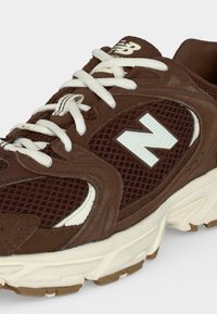 Καφέ αθλητικό παπούτσι New Balance με λευκά κορδόνια και λογότυπο, αναπνέον επάνω μέρος από δίχτυ και παχιά λευκή σόλα με καφέ πέλμα.