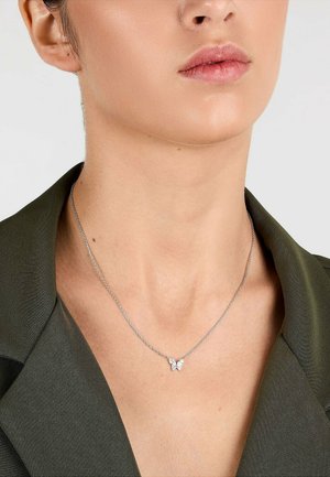 Nahaufnahme von Hals und Kiefer mit einer silbernen Kette und einem kleinen schmetterlingsförmigen Anhänger, bekleidet mit einem dunkelgrünen Blazer.