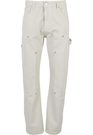 Homme portant un pantalon cargo écru avec des poches latérales et des baskets Nike vertes et blanches, debout devant un fond blanc uni.