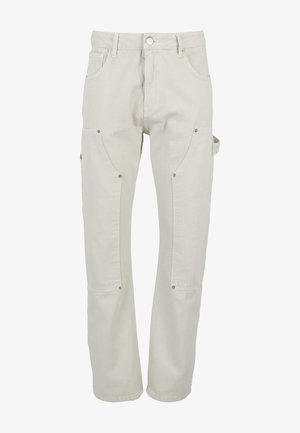 Homme portant un pantalon cargo écru avec des poches latérales et des baskets Nike vertes et blanches, debout devant un fond blanc uni.