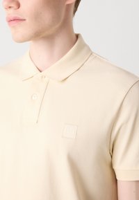 Polo majica u svijetloj bež boji, izrađena od teksturiranog materijala. Sadrži klasični ovratnik, dva dugmeta i mali četvrtasti logo u tonovima koji se slažu.