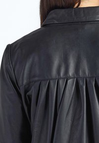 Sort læderjakke med en glat tekstur, der har et plisseret rygdesign, spids krave og rene syningsdetaljer.