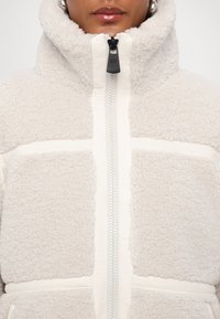 Fleece-takki korkealla kauluksella, beige väri, edessä vetoketju ja vaalean kermaiset yksityiskohdat. Tekstuuroitu kangas, jossa vaakasuuntainen paneelimuotoilu.
