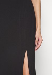 Robe noire avec une fente sur le côté, arrivant jusqu'à la cuisse, fabriquée dans un tissu doux et fluide. Main visible tenant la robe au niveau de la fente.