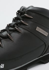 Stivale da trekking in pelle nera con occhielli metallici, finitura texturizzata e punta rinforzata. Presenta un'etichetta con logo argento e un design della suola robusta.