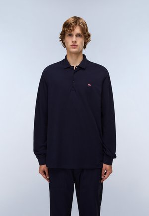 Młody mężczyzna o kręconych włosach, ubrany w granatową koszulkę polo z długim rękawem i pasujące spodnie, stojący na tle jasnoszarego tła.