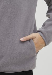 Pullover in fleece grigio caratterizzato da una texture morbida, tasca laterale e maniche lunghe con polsini aderenti. Design semplice senza motivi o dettagli decorativi.