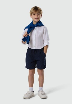 Jeune garçon portant une chemise blanche à manches longues, un short bleu marine, des baskets blanches et un pull bleu drapé sur les épaules, debout avec une main dans la poche.