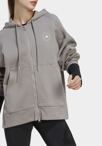 Sudadera gris con cremallera hecha de tela suave, con un logo en el pecho, dos bolsillos y detalles en negro en las mangas. Acabado texturizado con un corte relajado.