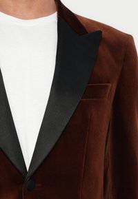 Bruine fluwelen blazer met zwarte satijnen revers, gedragen over een witte T-shirt met ronde hals, dichtgeknoopt aan de voorkant.