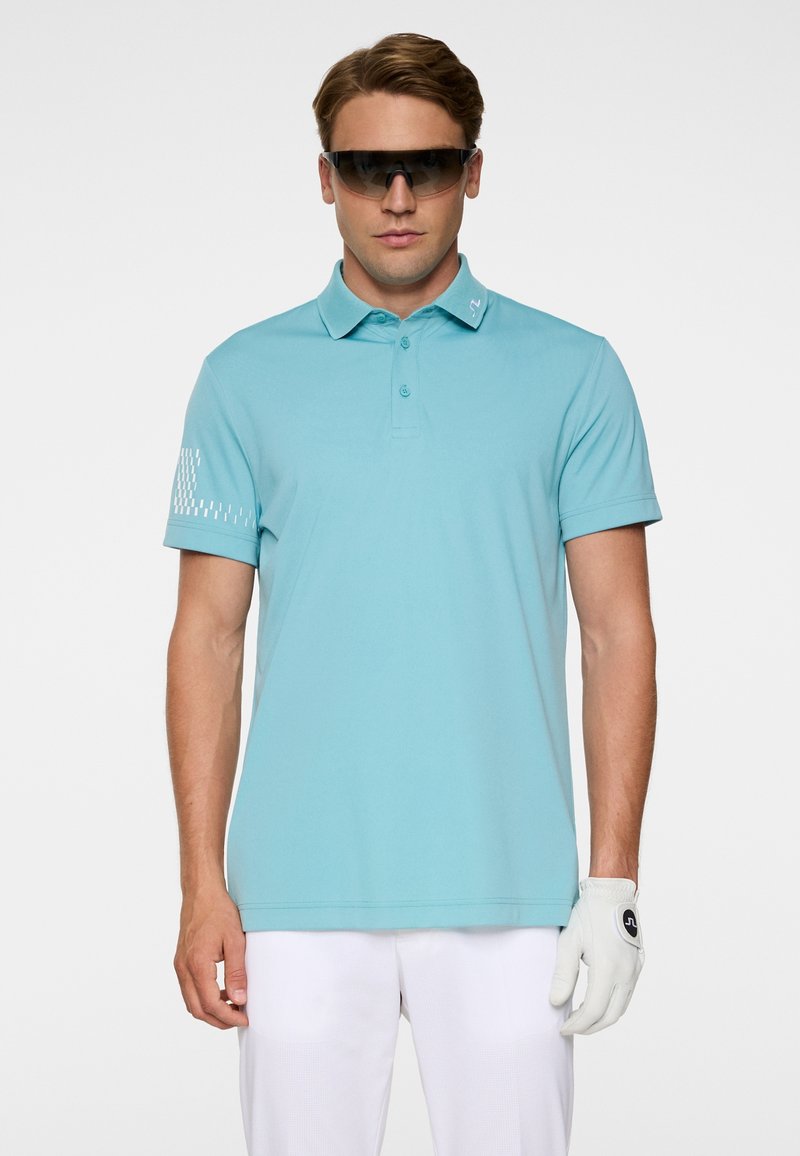 Homme portant un polo bleu clair, un gant de golf blanc à la main droite, un pantalon blanc et des lunettes de soleil foncées sur fond blanc.