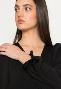 Blusa preta com mangas longas e franzidas. Material suave e leve, com decote em V e detalhes com babados nos punhos.