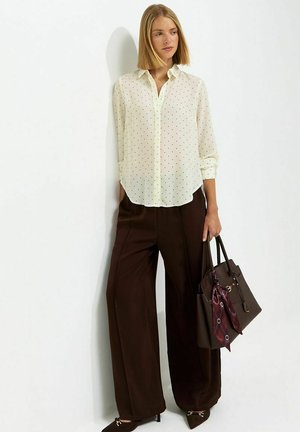 Mujer con una blusa blanca translúcida de lunares, pantalones anchos marrones, tacones marrones, sosteniendo un bolso marrón oscuro con un pañuelo rojo con estampado.
