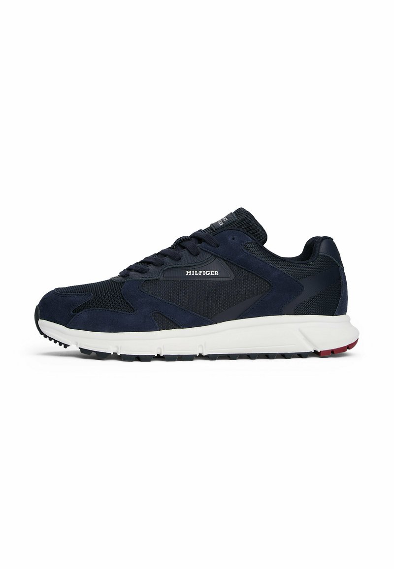 Zapatilla deportiva de color azul oscuro con paneles de malla negra y ante, suela blanca y logo "HILFIGER" en el lateral, mostrada desde el lado exterior.