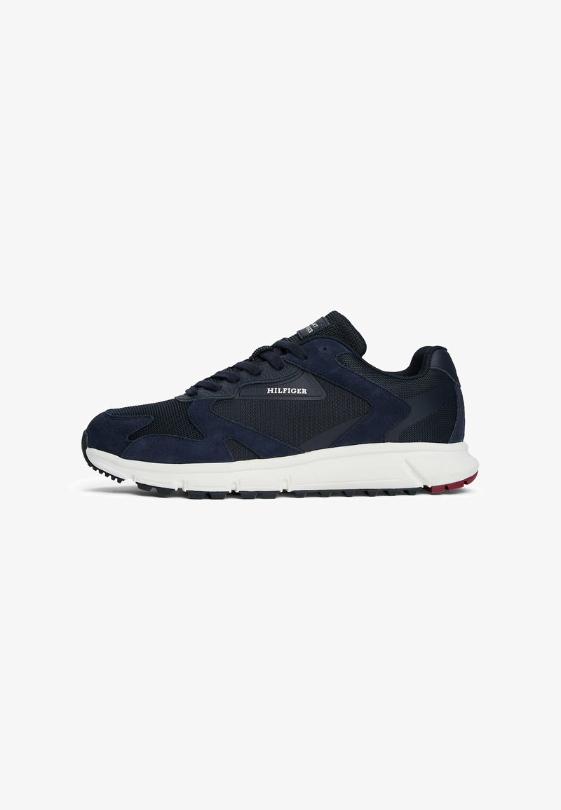 Donkerblauwe sport sneaker met zwarte mesh- en suedepanelen, witte zool en "HILFIGER" logo aan de zijkant, weergegeven vanaf de buitenkant.