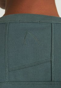 Tessuto di un verde scuro con texture, colletto a coste e cuciture visibili. Presenta un dettaglio triangolare ricamato sul pannello posteriore.