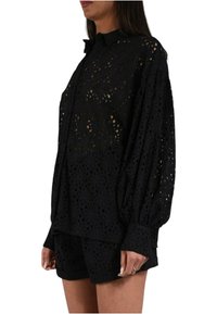 Blusa nera in pizzo con un motivo a fiori ritagliato, maniche lunghe e colletto con bottoni, abbinata a pantaloni corti neri coordinati.
