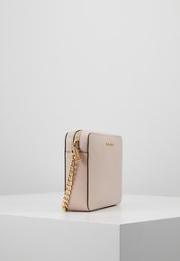 Kvadratisk, ljusrosa crossbody-väska med en texturerad yta, guld kedjesnöre och dragkedjestängning. Har en liten tagg och guldlogotyp på framsidan.