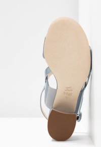 Sandale en cuir bleu clair avec une texture lisse, dotée d'une semelle en caoutchouc beige et d'une sangle élastique pour le confort.