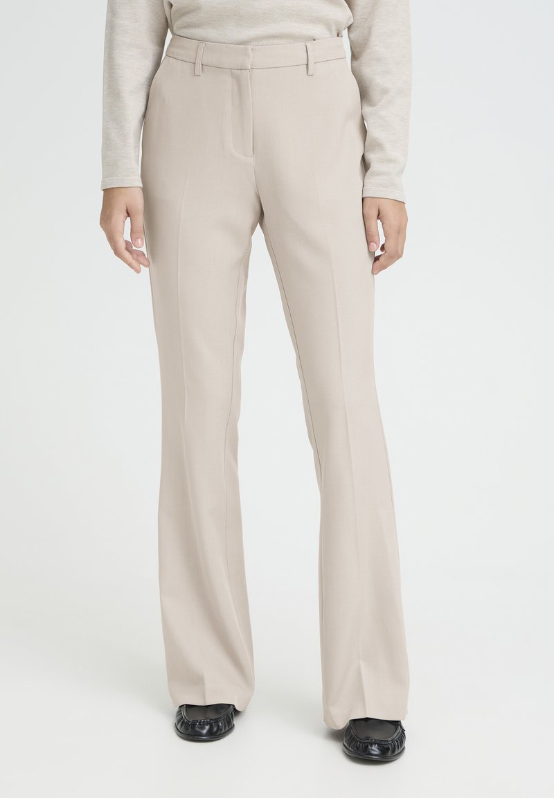Pantalon beige à jambes larges avec un design structuré, comprenant des plis à l'avant, des passants pour ceinture et une texture lisse. Assorti à des chaussures noires.