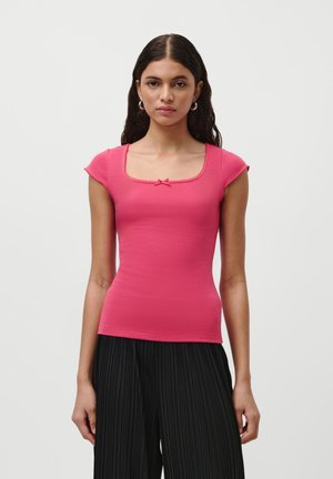 Haut rose ajusté à côtes avec de courtes manches à emmanchure, décolleté carré et un petit nœud ornemental à l'avant. Associé à un pantalon plissé noir.