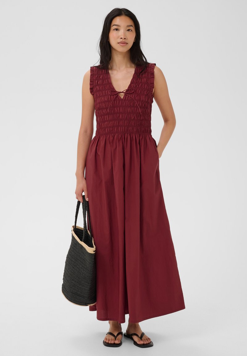 Femme portant une robe maxi bordeaux sans manches avec un corsage smocké, tenant un sac cabas noir tissé et portant des sandales noires, debout devant un fond blanc.