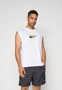Rövid sötét hajú férfi fehér ujjatlan Nike pólóban és fekete Nike sortban áll egy sima háttér előtt.
