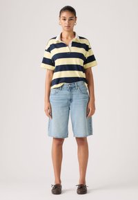 Γυναίκα με στεφάνια σε σκουλαρίκια, φορώντας μπλε ναυτικό και κίτρινο ριγωτό πόλο πουκάμισο, ανοιχτόχρωμα τζιν σορτς μέχρι το γόνατο και καφέ δερμάτινα παπούτσια με κορδόνια.