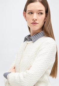 Femme aux longs cheveux bruns portant un cardigan blanc en tricot torsadé par-dessus une chemise rayée à col, regardant sur le côté les bras croisés.