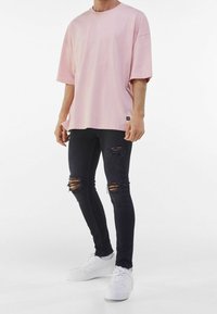 Muž v nadměrné růžové tričko, roztrhaných černých skinny džínách s dírami na kolenou a bílých teniskách na jednotném pozadí.