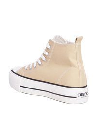 Zapatillas altas de lona beige con cordones blancos, puntera de goma y una suela blanca gruesa con una franja negra y la marca en el talón.
