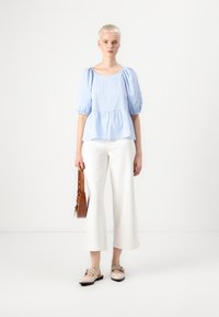 Lyseblå rutete peplum-bluse med puffede ermer, kombinert med hvite vide bukser og beige sko, bærende en brun crossover-veske.