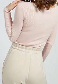 Žena v proužkovaném světlém růžovém triku s dlouhým rukávem a v béžových kalhotách s vysokým pasem, pózující s jednou rukou u ramene.