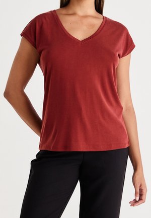 Kobieta ubrana w rdzawy, krótki rękaw, koszulkę z dekoltem w serek i czarne spodnie, stoi z jedną ręką za plecami.