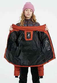 Orange isoleret jakke med sort indvendig, der har mærker og en meshlomme. Båret over en farverig mønstret top og sorte bukser.