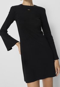 Černé šaty s dlouhými rukávy, ribbovanou texturou a kulatým výstřihem. Mají rozšířené okraje rukávů a malý zlatý kovový detail.