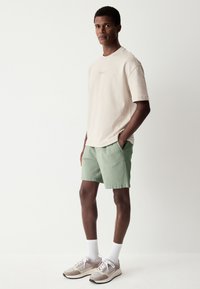 T-shirt beige texturé, short vert, chaussettes blanches et baskets gris-brun. Le modèle est debout, les mains dans les poches, sur un fond clair.