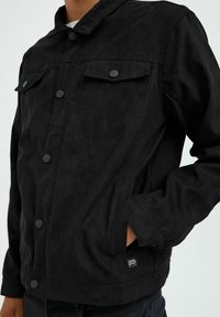 Veste en suede noire avec des boutons-pression, deux poches avant et une étiquette près de l'ourlet. Elle présente un col et une coupe décontractée.