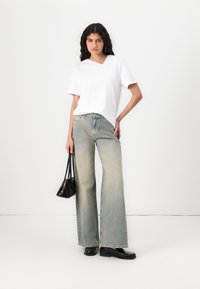 T-shirt bianco con scollo a V, jeans larghi blu chiaro e ballerine nere. Il modello tiene una piccola borsa nera. Tessuto morbido, vestibilità casual, design minimal.