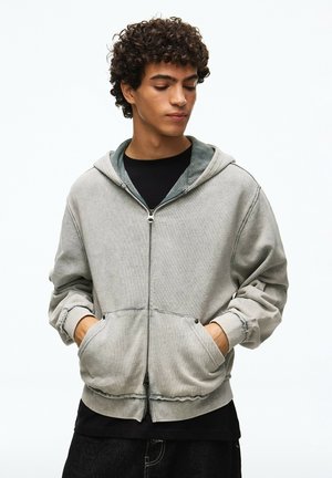 Junger Mann mit lockigem Haar, der eine hellgraue Kapuzenjacke mit Reißverschluss trägt und die Hände in den Taschen hat, schaut leicht nach unten und zur Seite.