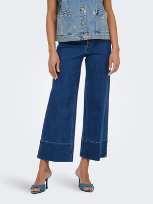 Modèle portant un jean bleu large et court associé à un gilet en denim sans manches et des sandales à talons ouvertes bleues.