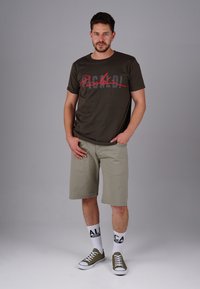 Olivgrünes T-Shirt mit rotem Schriftzug-Graphic, beige Shorts, weiße Socken mit schwarzem Text und grüne Sneaker. Model steht vor einem grauen Hintergrund.