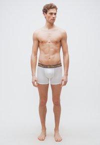 Νέος άνδρας στέκεται ξυπόλητος φορώντας λευκά εσώρουχα τύπου μποξεράκια με ζώνη μέσης σε μωβ και χρυσό μοτίβο, σε απλό λευκό φόντο.