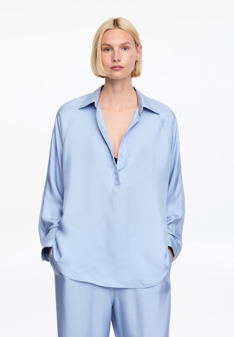 Femme aux cheveux blonds courts portant un chemisier ample en satin bleu clair et un pantalon large assorti, mains dans les poches, expression neutre.
