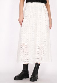 Jupe midi blanche en tissu brodé avec des motifs découpés. Elle présente une ceinture à nouer à la taille et est associée à des bottes noires à la cheville.